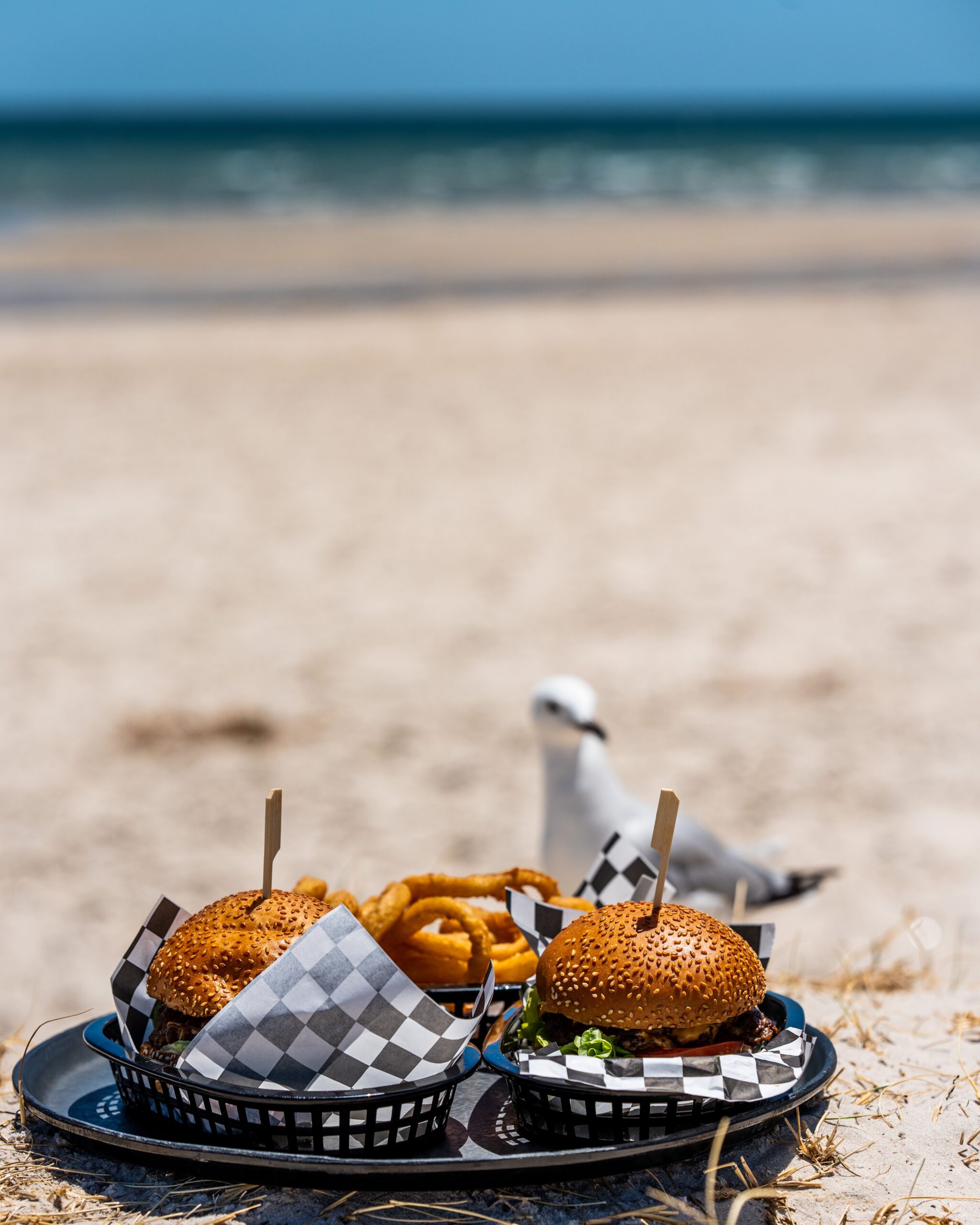 Two baskets of burgers and onion rings on the beach with a seagull photobombing