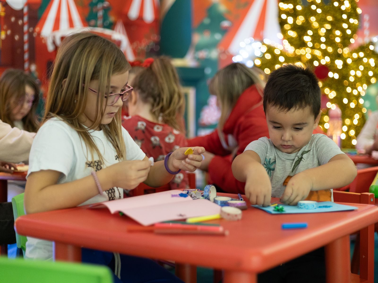 Children making holiday crafts at a red table in a festive decorated room
