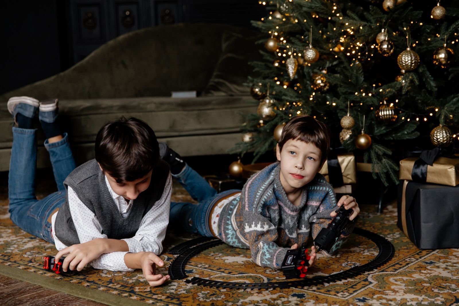 Two boys lying on a rug playing with a toy train next to a Christmas tree